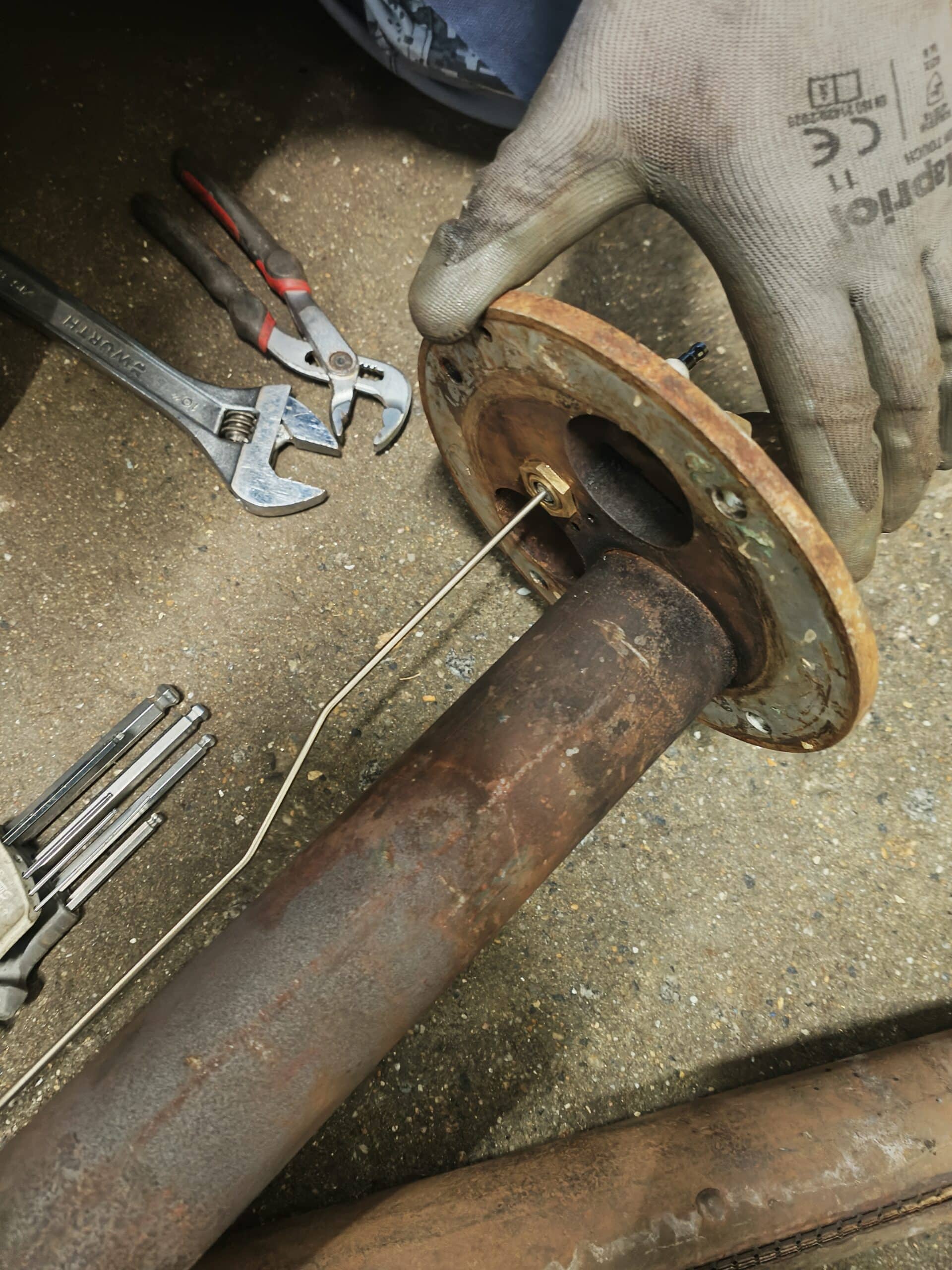 Main gantée inspectant une bride métallique rouillée avec clé Wurth, pince et clés Allen sur béton.