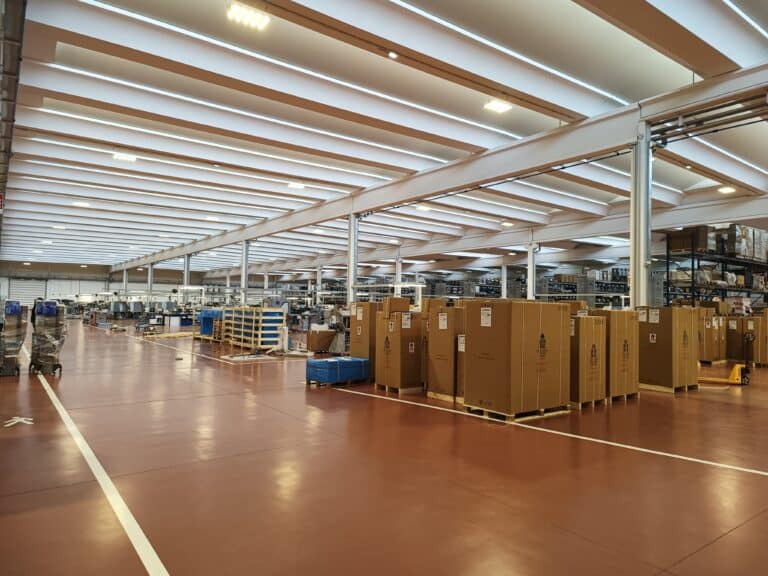 Entrepôt logistique moderne avec cartons sur palettes, sol rouge brillant et structure de toit industrielle lumineuse.